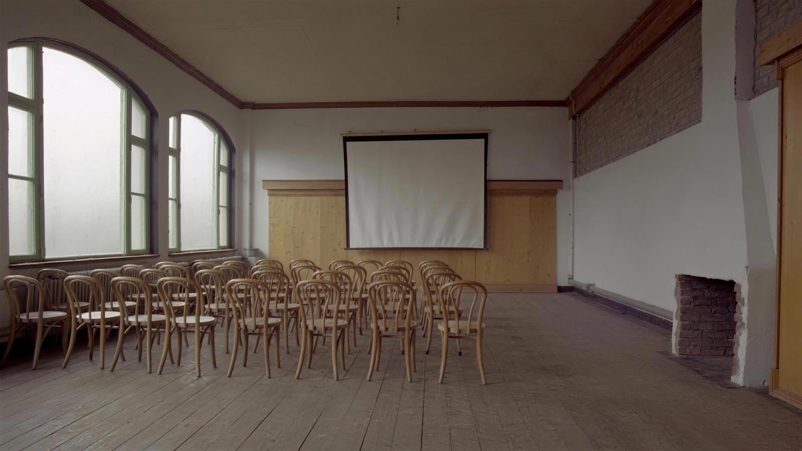Bestuhlter Saal mit Holzstühlen in Reihen vor einer weißen Leinwand, Tageslicht durch hohe Sprossenfenster